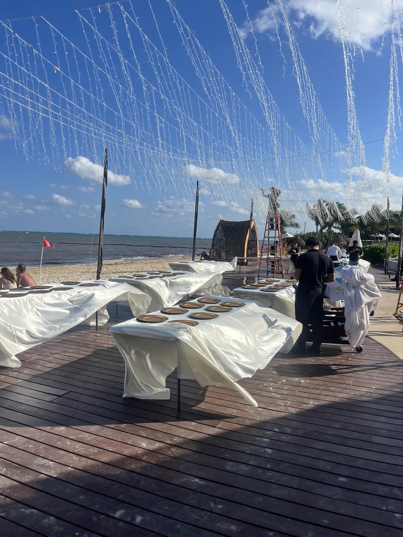 Long banquet tables set up on the beach at sunset with string lights overhead